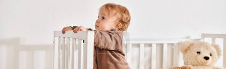 curious little boy standing in crib with teddy bear in cozy nursery room at home, horizontal banner