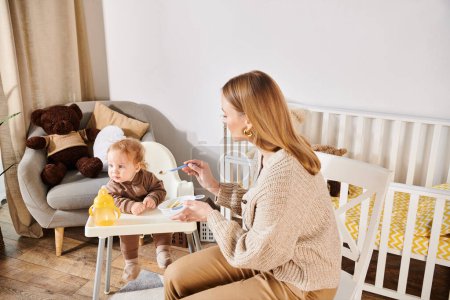 Foto de Mujer joven alimentando a su hijo pequeño con desayuno en silla de bebé en la habitación de la guardería, crianza moderna - Imagen libre de derechos