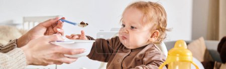 Foto de Mujer joven alimentando a un niño pequeño con desayuno en silla de bebé en la habitación de la guardería, pancarta horizontal - Imagen libre de derechos