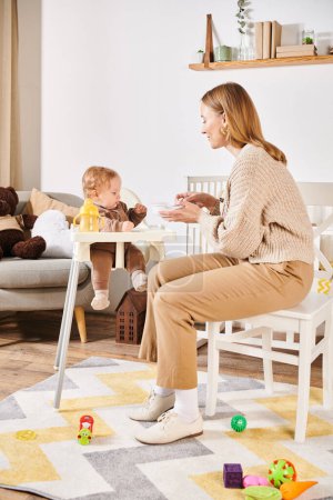 Foto de Feliz mujer alimentación niño pequeño hijo con desayuno en silla de bebé en la habitación de la guardería, maternidad dichosa - Imagen libre de derechos