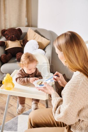 madre joven alimentando a su hijo pequeño con el desayuno en la silla de bebé en la habitación de la guardería, maternidad dichosa