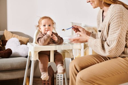 smiling woman feeding toddler boy with breakfast on baby chair in nursery room, blissful motherhood