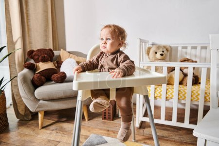 Foto de Niño pequeño sentado en silla de bebé y mirando hacia otro lado en la acogedora habitación del vivero con cuna y osos de peluche - Imagen libre de derechos