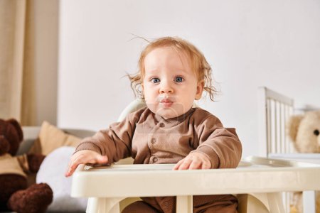 Foto de Retrato de lindo niño sentado en silla de bebé y mirando a la cámara en la habitación de la guardería en casa - Imagen libre de derechos