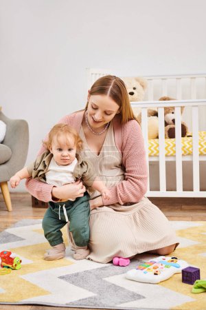 smiling woman supporting toddler son learning to walk on floor in living room at home, motherhood