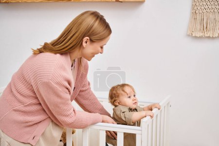 Foto de Sonriente mujer de apoyo hijo pequeño de pie en la cuna en la habitación de la guardería en casa, la maternidad feliz - Imagen libre de derechos
