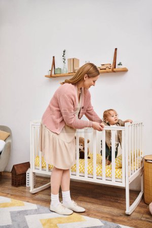 Foto de Alegre mujer de apoyo pequeño hijo de pie en la cuna en la habitación de la guardería en casa, la maternidad feliz - Imagen libre de derechos