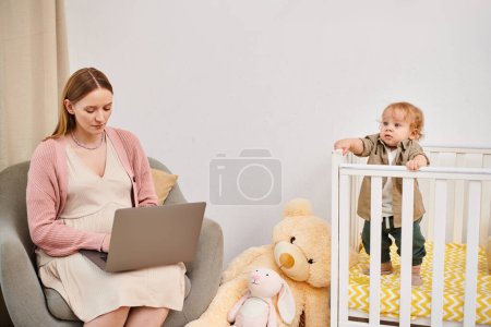 Foto de Mujer embarazada que trabaja en el ordenador portátil cerca de su hijo pequeño en la cuna en la habitación de la guardería, multitarea - Imagen libre de derechos