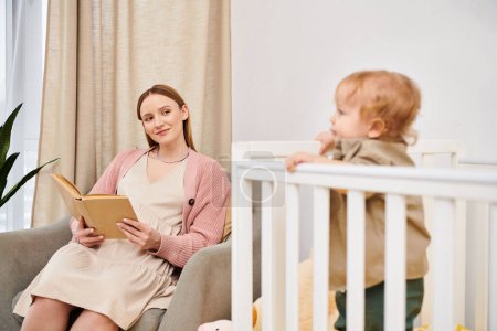 Foto de Feliz embarazada lectura libro cerca adorable niño hijo en cuarto de la guardería en casa, maternidad - Imagen libre de derechos