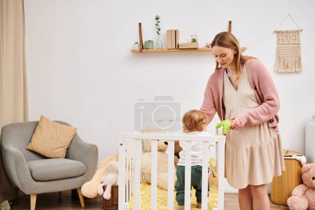 Foto de Sonriendo mujer embarazada sosteniendo juguete y jugando con su hijo pequeño de pie en la cuna en la habitación de la guardería - Imagen libre de derechos