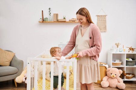 Foto de Alegre embarazada celebración juguete mientras jugando con pequeño hijo de pie en cuna en habitación de la guardería - Imagen libre de derechos