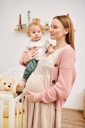 jeune femme enceinte enchantée debout avec tout-petit fils dans la chambre d'enfant à la maison, maternité