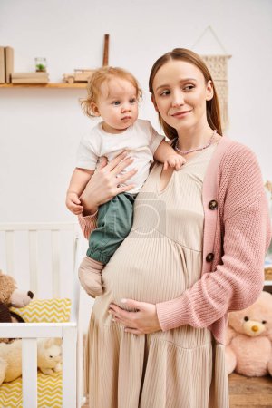 pleased young pregnant woman standing with toddler son in nursery room at home, happy motherhood