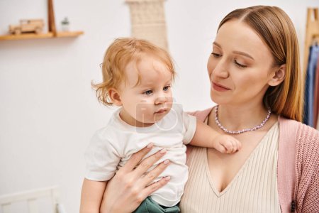 fröhliche attraktive Frau mit kleinem Kleinkindsohn in den Händen im Kinderzimmer, glückliche Mutterschaft