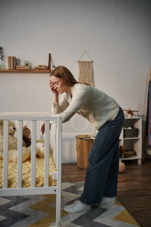 Frustrierte Frau steht neben Krippe mit Plüschtieren in trostlosem Kinderzimmer zu Hause, unglücklich