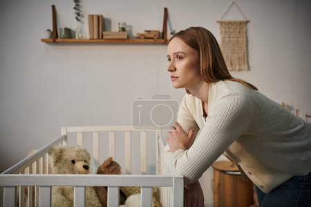 mujer descorazonada de pie cerca de la cuna con juguetes blandos en el cuarto oscuro de la guardería en casa, afligida