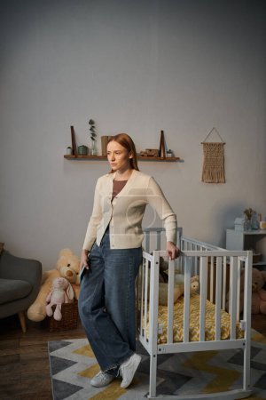 Foto de Mujer infeliz joven de pie cerca de la cuna con juguetes suaves en la habitación de la guardería sombría en el hogar, depresión - Imagen libre de derechos