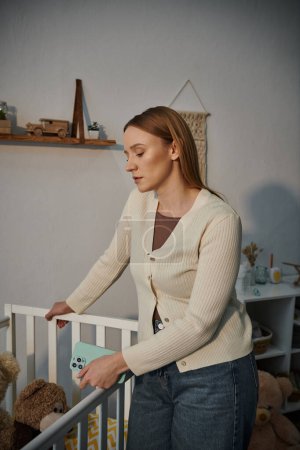Trauernde junge Frau mit Smartphone steht neben Krippe mit Plüschtieren im trostlosen Kinderzimmer zu Hause
