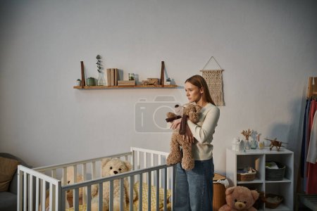 Hilflose und aufgebrachte Frau mit Stofftier steht neben Krippe in trostlosem Kinderzimmer zu Hause