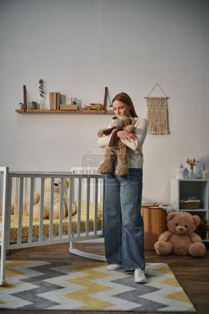 Frustrierte mutlose Frau mit Stofftier steht neben Krippe in trostlosem Kinderzimmer zu Hause