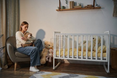 despairing young woman with soft toy sitting in armchair near crib in dark nursery room at home