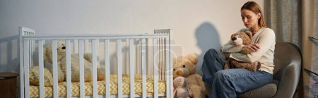 Photo for Depressed young woman with soft toy sitting in armchair near crib in dark nursery room, banner - Royalty Free Image