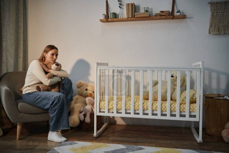 Hoffnungslose junge Frau mit Stofftier sitzt in Sessel neben Krippe mit in trostlosem Kinderzimmer zu Hause