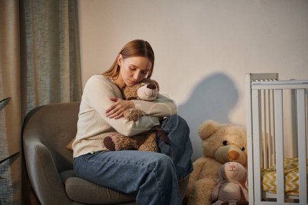 Trauernde tränenreiche Frau mit Stofftier sitzt im Sessel neben Krippe mit in trostlosem Kinderzimmer zu Hause