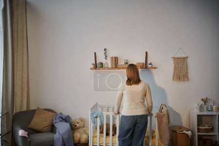Rückansicht einer depressiven und einsamen Frau in der Nähe von Krippe mit Plüschtieren und dunklem Kinderzimmer zu Hause