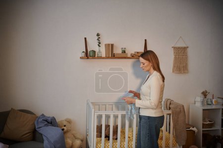 Téléchargez les photos : Vue latérale d'une femme frustrée tenant des vêtements de bébé près d'un berceau avec des jouets mous dans une chambre d'enfant - en image libre de droit