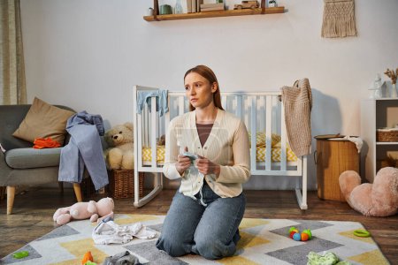 Foto de Mujer angustiada con ropa de bebé sentada en el suelo cerca de la cuna y juguetes en la habitación de la guardería en casa - Imagen libre de derechos