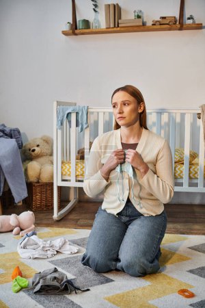 femme découragée avec des vêtements de bébé assis sur le sol près de la crèche et des jouets dans la chambre d'enfant à la maison