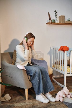 Foto de Mujer frustrada tratando de relajarse escuchando música en los auriculares en el cuarto oscuro de la guardería en casa - Imagen libre de derechos
