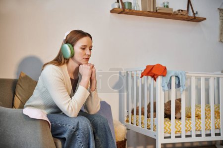 Foto de Mujer de corazón pesado tratando de relajarse escuchando música en los auriculares en el cuarto oscuro de la guardería en casa - Imagen libre de derechos