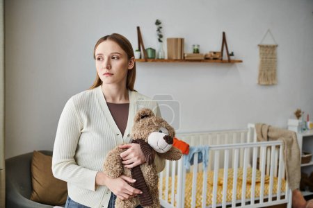 Téléchargez les photos : Jeune femme déprimée avec jouet doux regardant loin dans la chambre d'enfant à la maison, moment déchiré - en image libre de droit