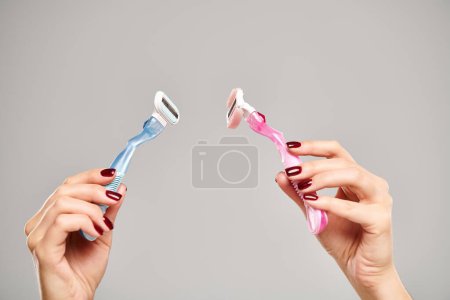 unknown young woman with nail polish holding blue and pink razors in her hands on gray background