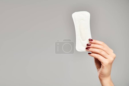 white sanitary pad in hand of unknown female model with nail polish on gray backdrop, hygiene