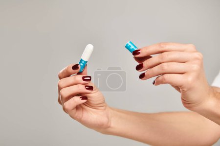 hygienic tampon in hands of unknown young female with nail polish on gray backdrop, object photo