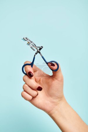 object photo of eyelash curler in hand of young unknown woman with nail polish on blue backdrop