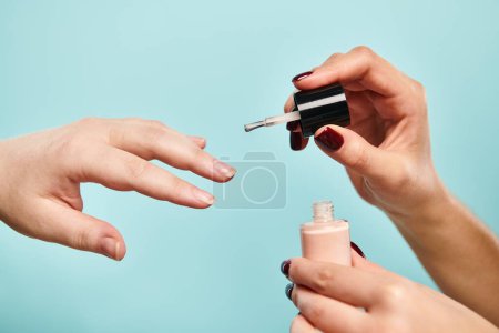 young unknown woman covering nails of other person with beige polish on vibrant blue background