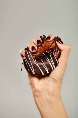 unknown young woman with nail polish squeezing tasty donut with brown icing on gray background