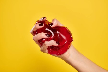 Foto de Mujer joven desconocida con esmalte de uñas exprimiendo gelatina roja deliciosa en su mano sobre fondo amarillo - Imagen libre de derechos
