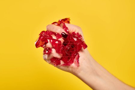 mujer joven desconocida con esmalte de uñas apretando gelatina roja fresca en su mano sobre fondo amarillo vivo