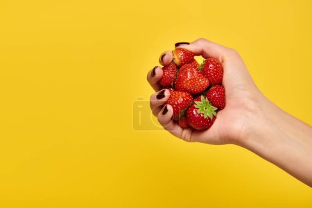 object photo of red fresh delicious strawberries in hand of young unknown woman on yellow backdrop