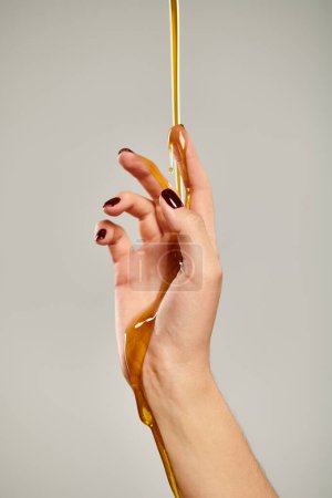 hand of unknown young woman with nail polish covered with sweet fresh honey on gray backdrop