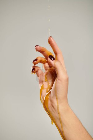 hands of unknown woman with dark nail polish covered with organic golden honey on gray backdrop