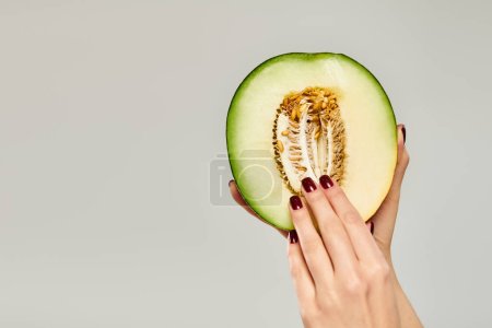 mujer joven desconocida con esmalte de uñas recogiendo semillas de melón fresco sobre fondo gris