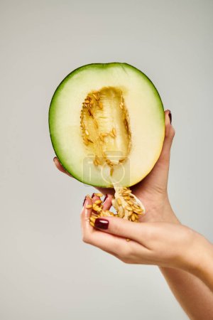 mujer desconocida con esmalte de uñas recogiendo semillas de delicioso melón sobre fondo gris