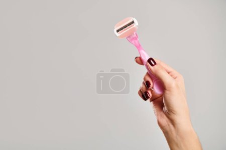 Objektfoto von rosa Rasiermesser in der Hand eines unbekannten weiblichen Modells mit Nagellack auf grauem Hintergrund