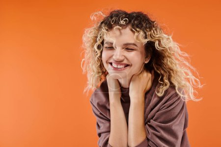 trendy woman with wavy hair in mocha color turtleneck smiling with closed eyes on orange backdrop